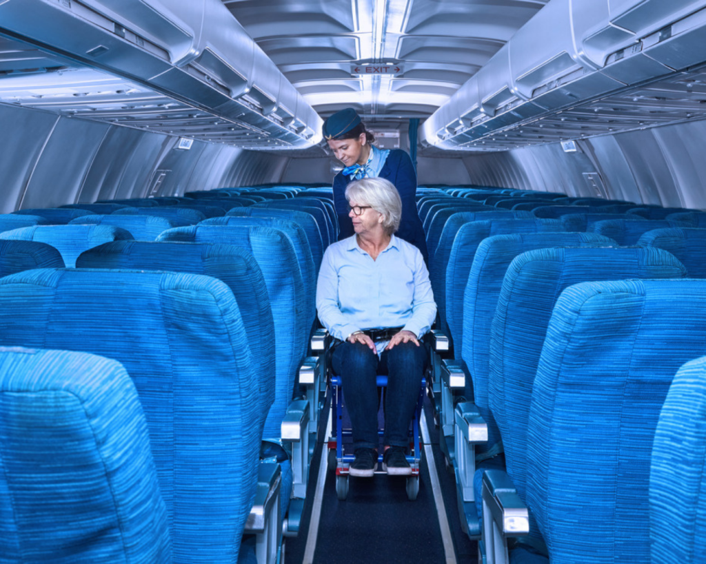 Passenger seated in an Airchair II onboard wheelchair being assisted down the aircraft aisle by a flight attendant.